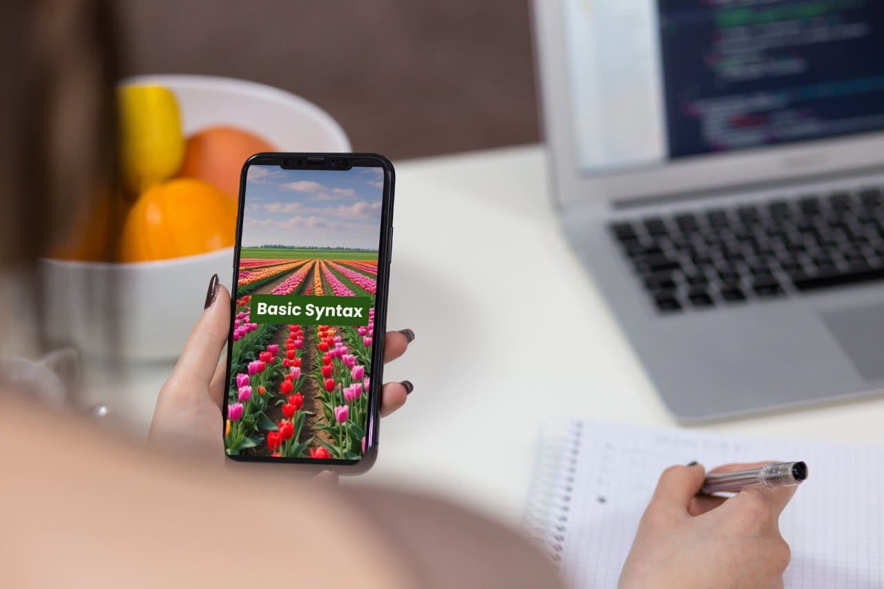 Person holding a phone with a screen displaying 'Basic Syntax' C++ course cover over a field of tulips, with a laptop and fruit bowl in the background.